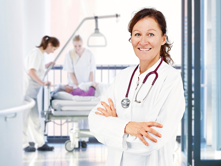 Werden Sie Ärzt*in bei KMG Kliniken! Ärztin lächelt, Im Hintergrund Patient*in mit Pflegekräften